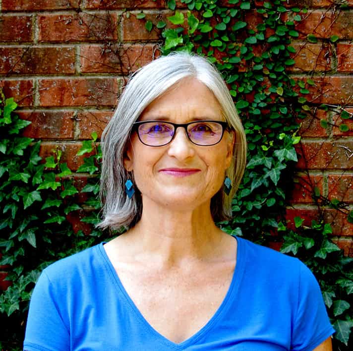 Portrait of a woman with gray hair and glasses, standing in front of a brick wall with green ivy.