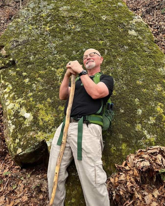 Moss gardening enthusiast leaning on a wooden staff in front of a large moss-covered rock.
